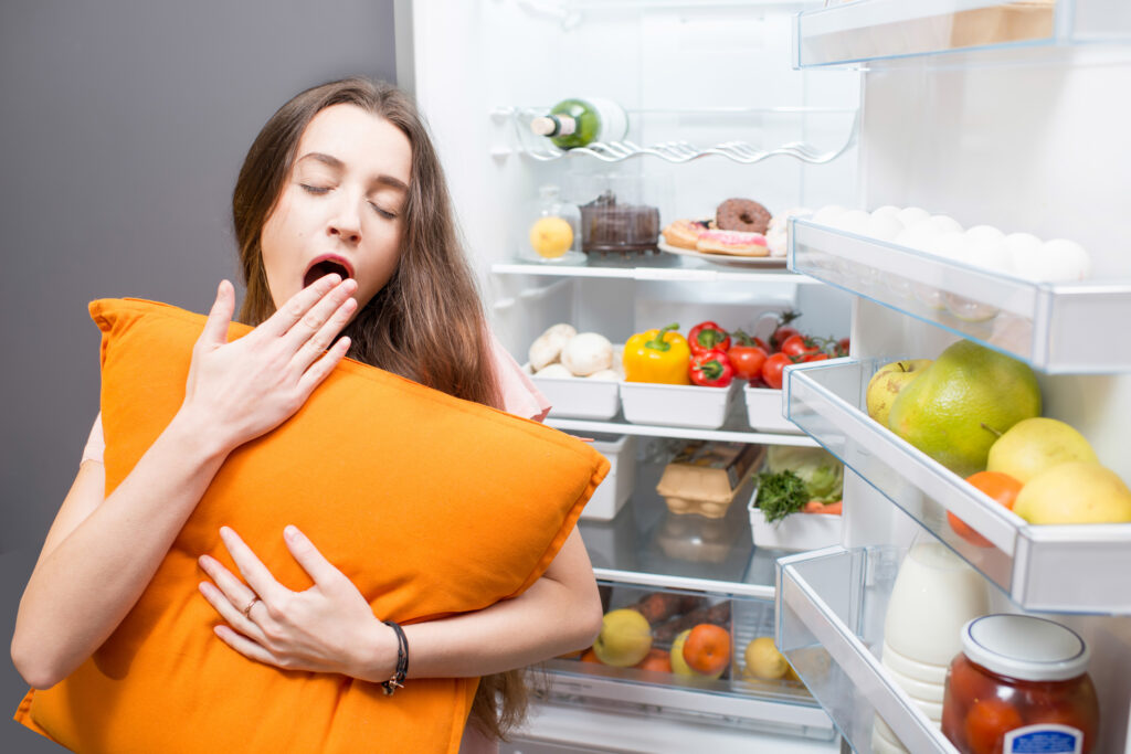 mulher com sono em frente a geladeira, segurando travesseiro amarelo