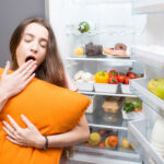 mulher com sono em frente a geladeira, segurando travesseiro amarelo